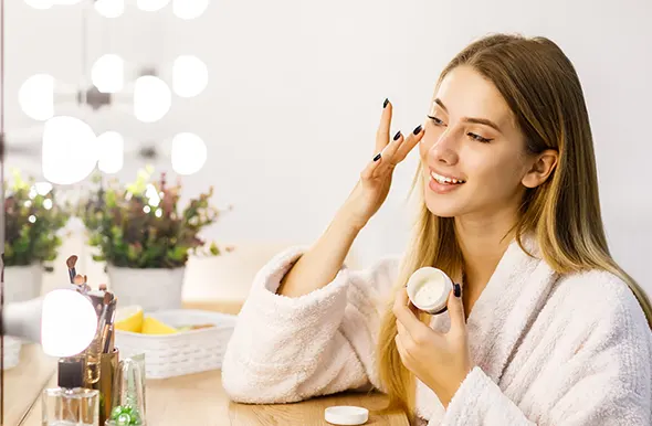 A smiling woman in a robe applying face cream in front of a bright vanity mirror.