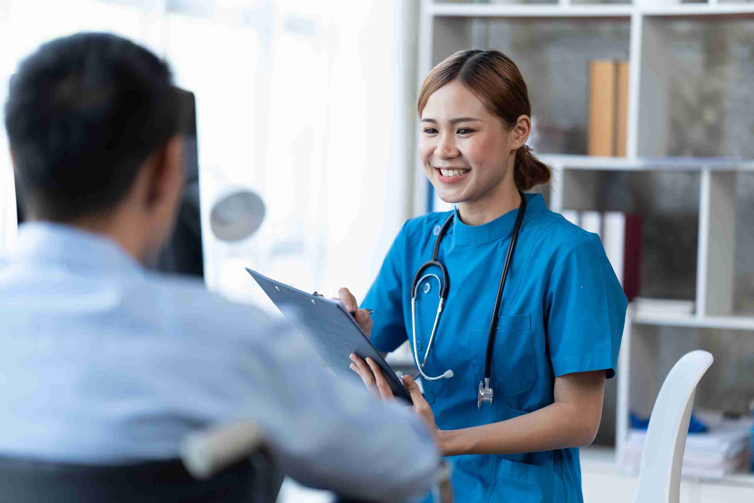 A smiling healthcare provider in blue scrubs holds a clipboard while talking to a patient.