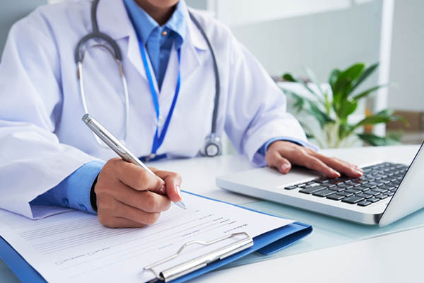 A doctor’s hands writing on a clipboard while using a laptop in a bright medical office.