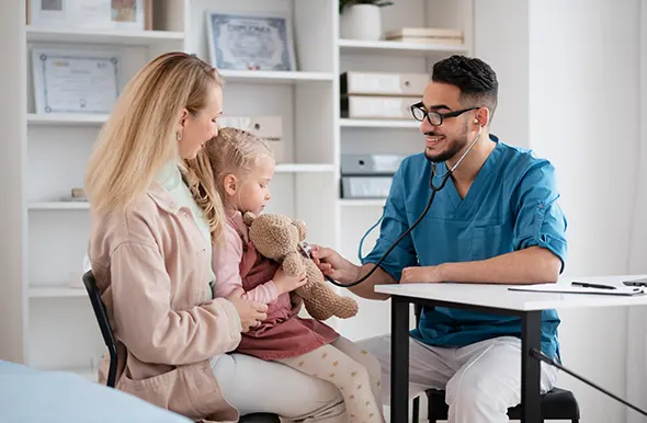 A male doctor in blue scrubs uses a stethoscope to examine a young girl's teddy bear.